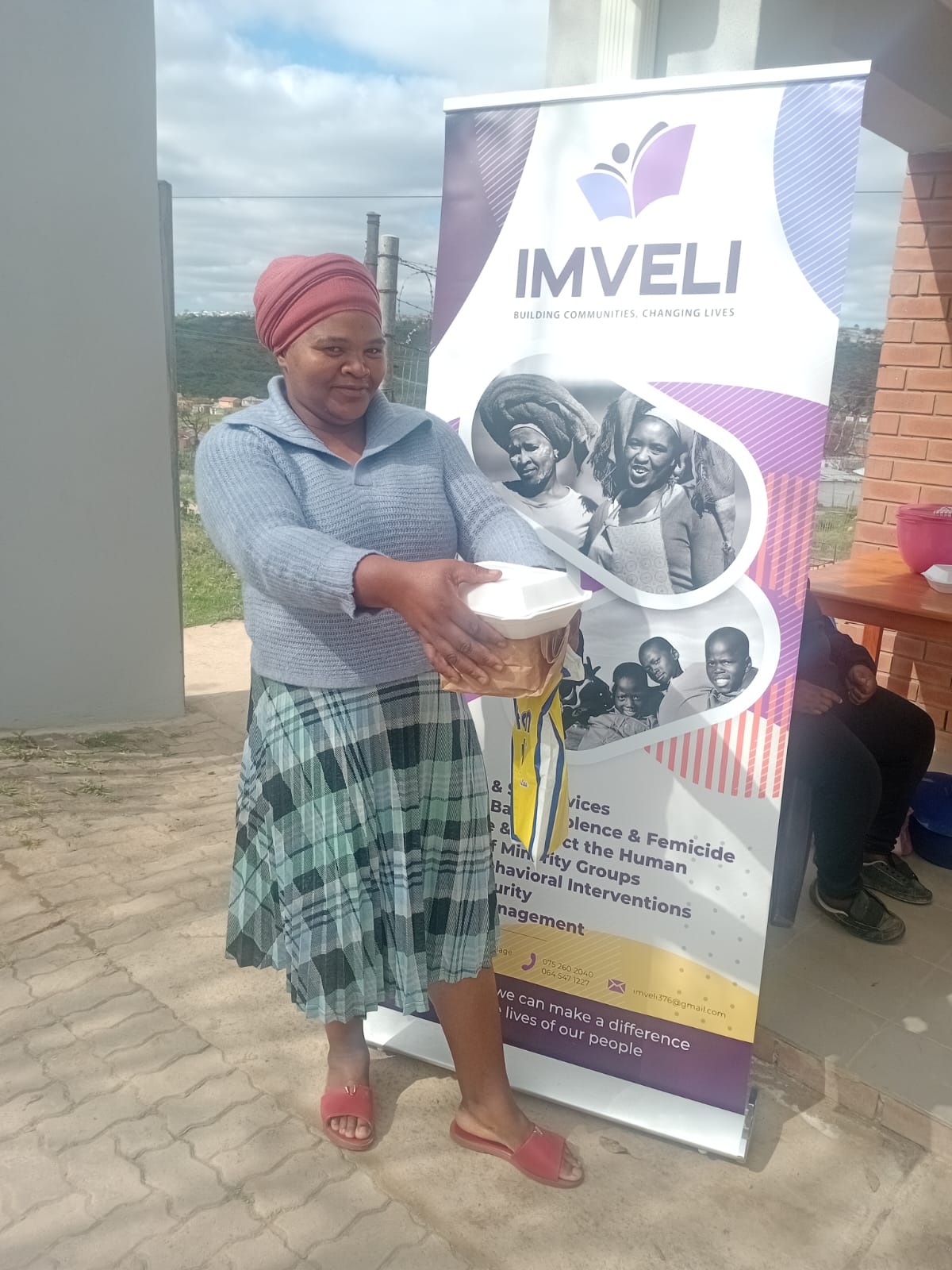 A woman gratefully receives a hot meal container beside an Imveli banner