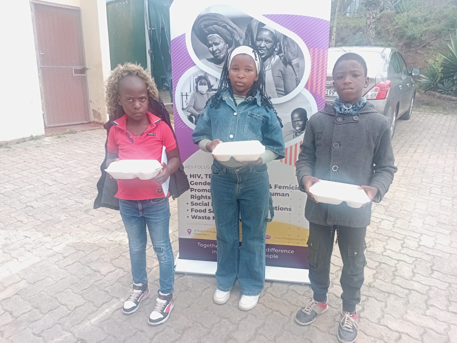 Three children hold meal containers in front of an Imveli banner at a community meal distribution