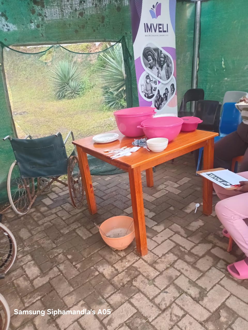 A wooden table with pink meal containers, plates and utensils set out for community meal service, with an Imveli banner and participants nearby