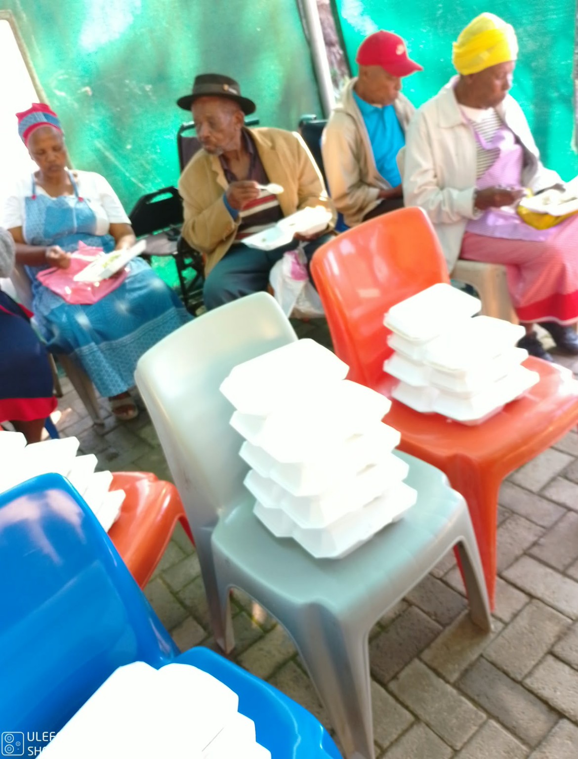 Stacks of prepared meal containers on chairs while community members seated nearby eat their meals