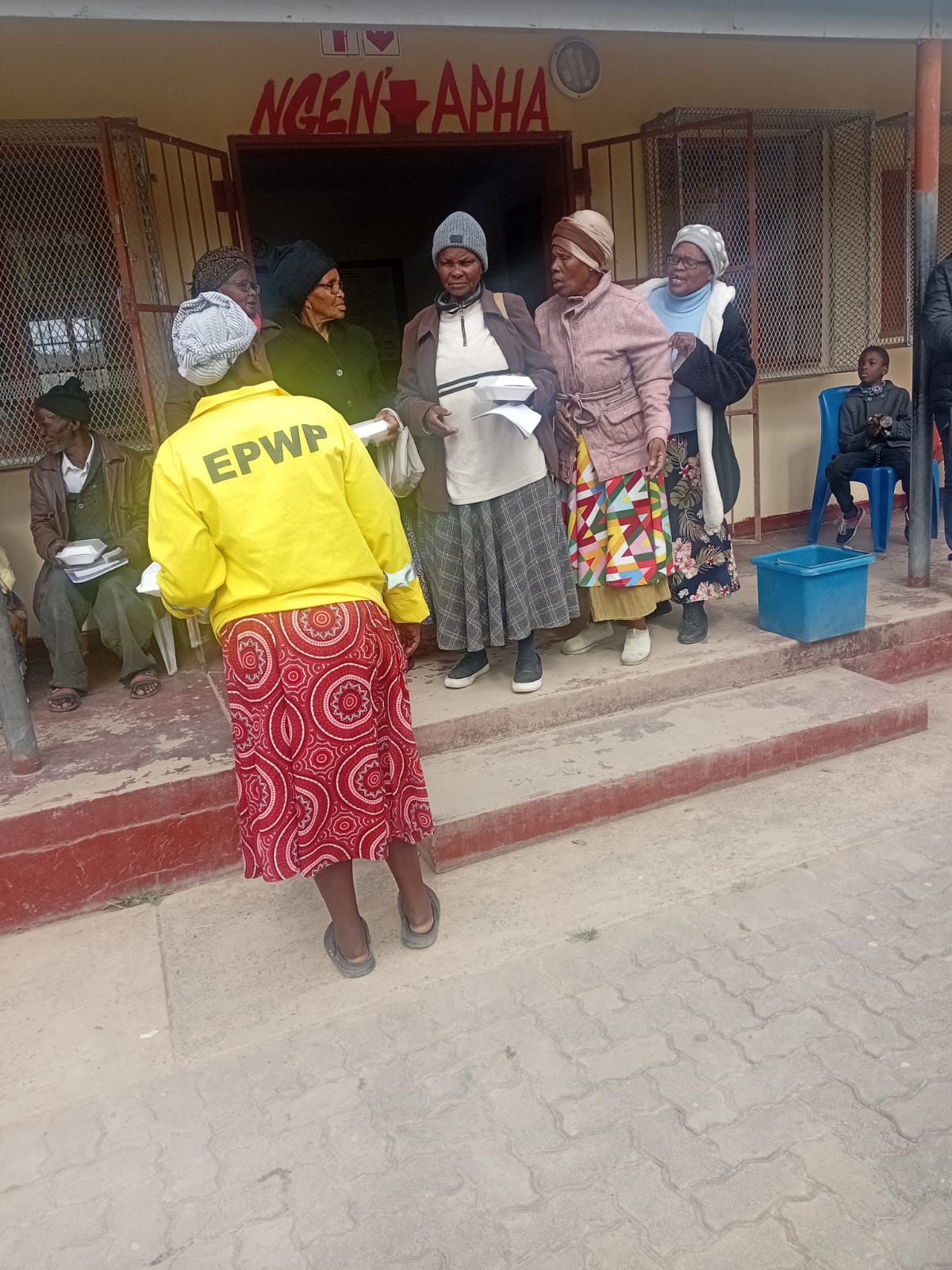 An EPWP volunteer hands meals to elderly community members waiting outside Potsdam Clinic