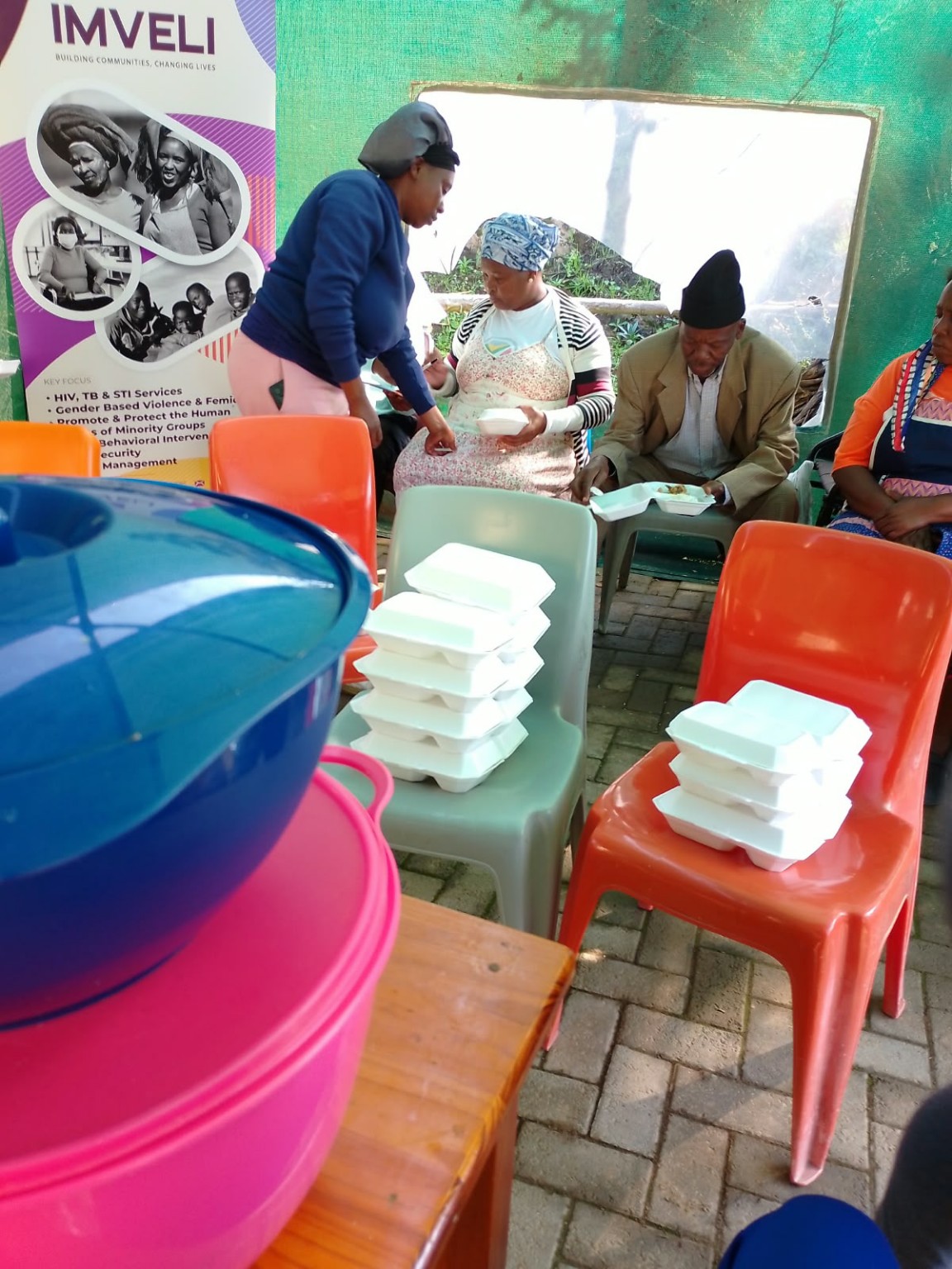 A volunteer handing out prepared meal containers to seated community members next to an Imveli branded banner