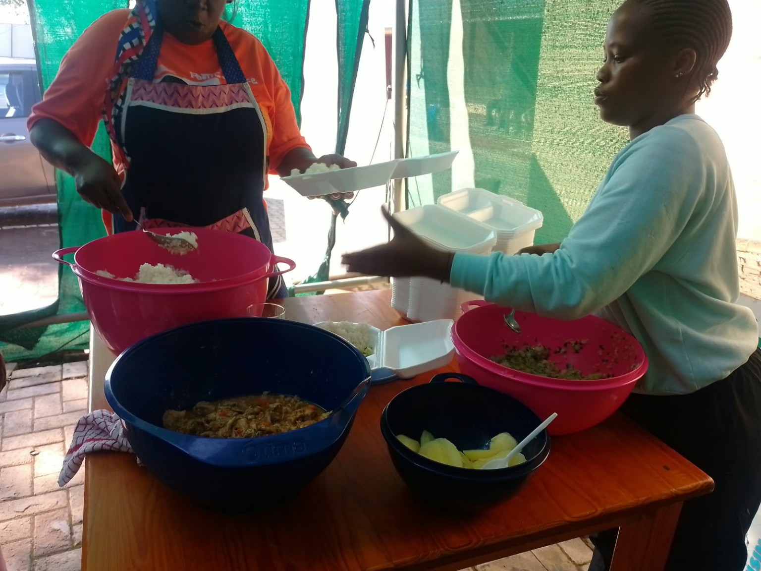 Two community volunteers serving rice and a cooked dish into takeaway meal containers at a preparation table