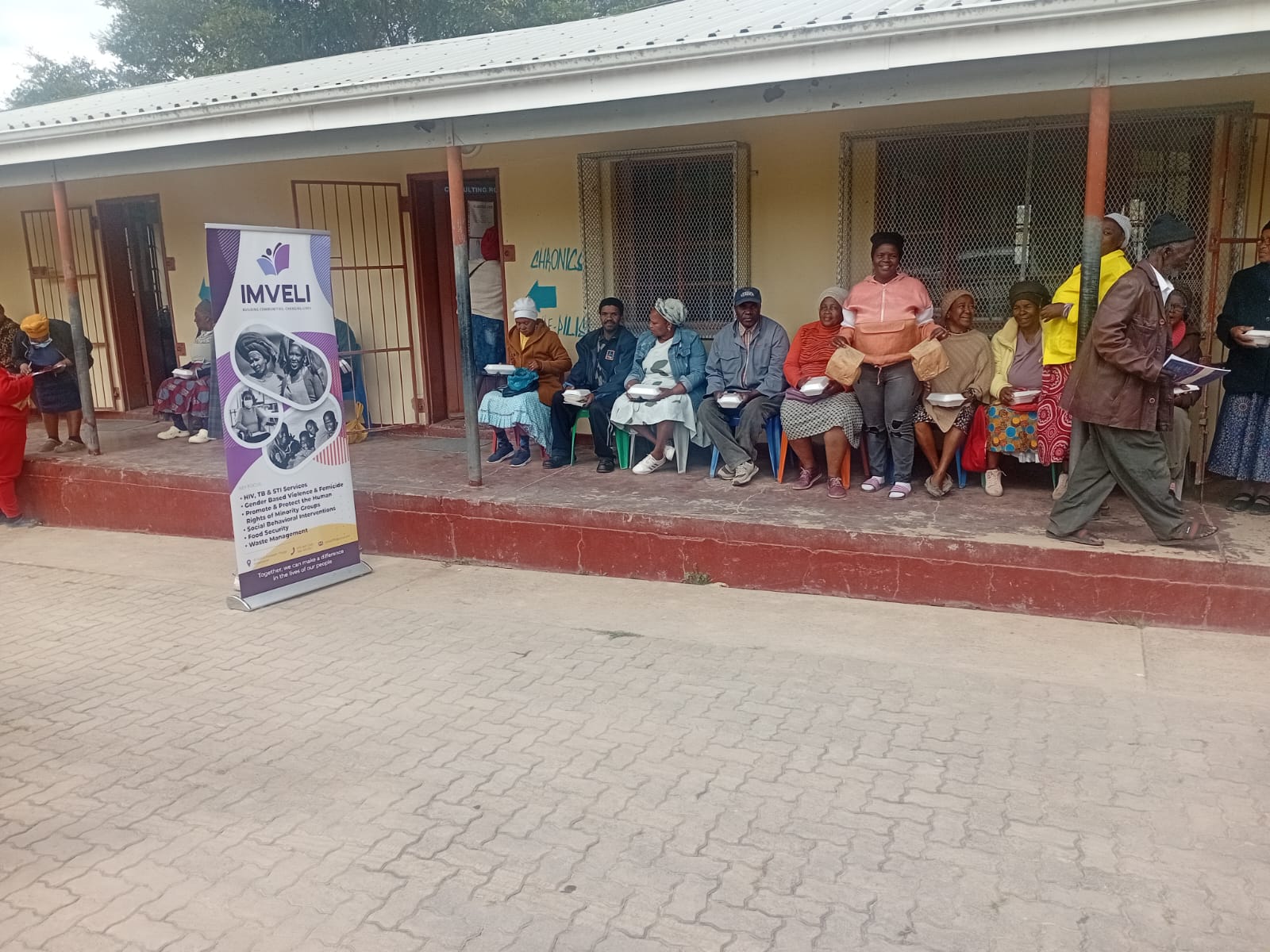 Community members seated outside Potsdam Clinic holding meal containers during a One Meal a Day distribution